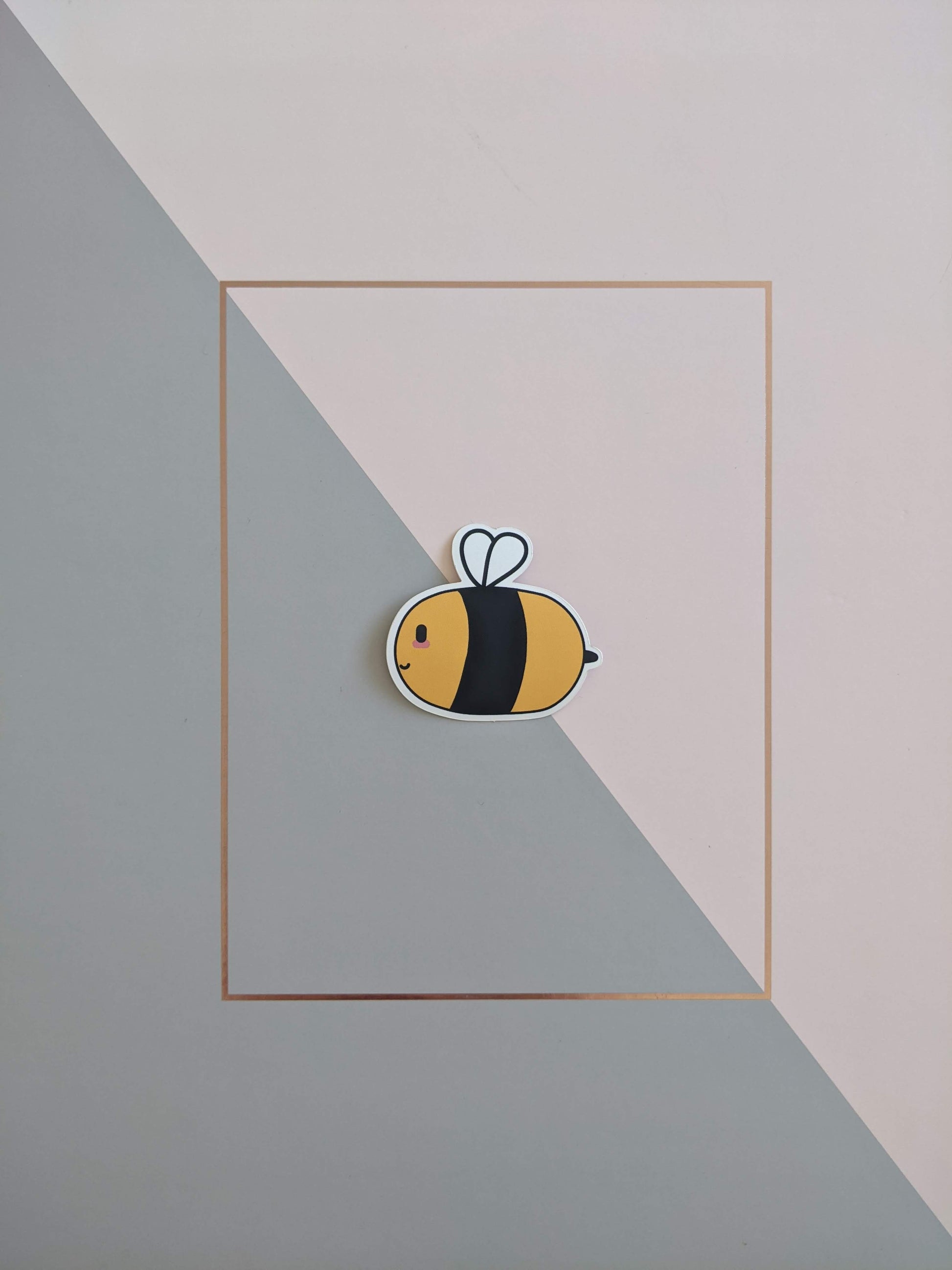 A bumblebee sticker on a pink and grey background. The bee has a smiley face and blushed cheeks, it has a chunky black and yellow body with small white wings.