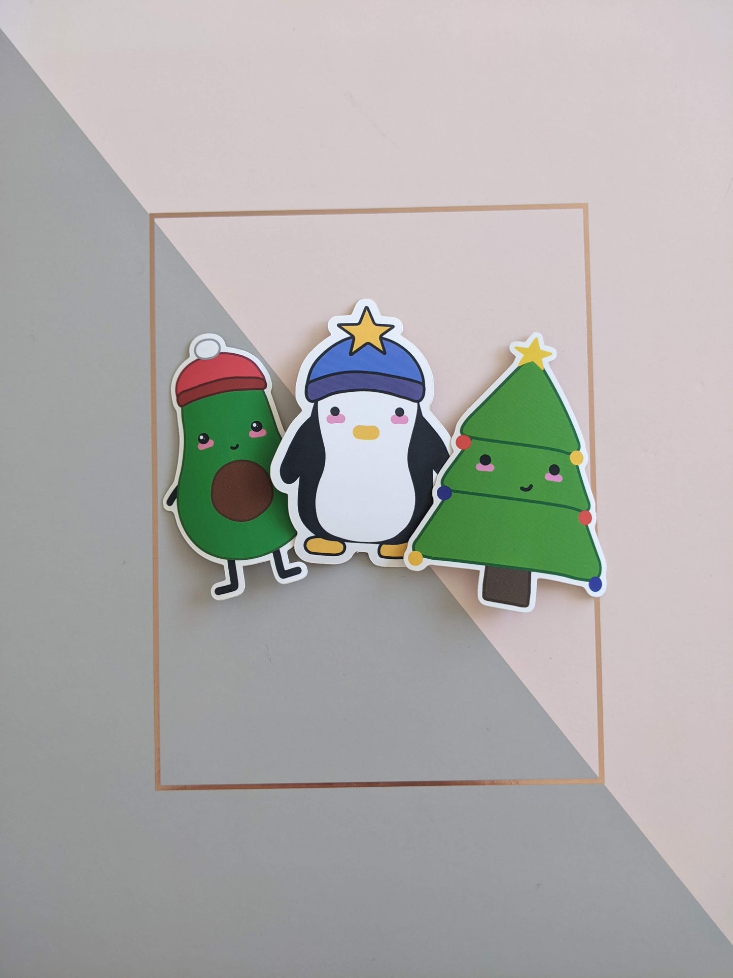 3 stickers on a pink and grey background. On the left is a halved avocado with a smiley face and blushed cheeks, wearing a red beanie hat with a white pompom on top. In the centre is a chunky penguin with a blank expression and blushed cheeks, wearing a blue beanie hat with a yellow star on top. On the right is a Christmas tree with a cheeky smile and blushed cheeks, he has 6 baubles, 2 red, 2 royal blue and 2 yellow, as well as a yellow star at the top. 