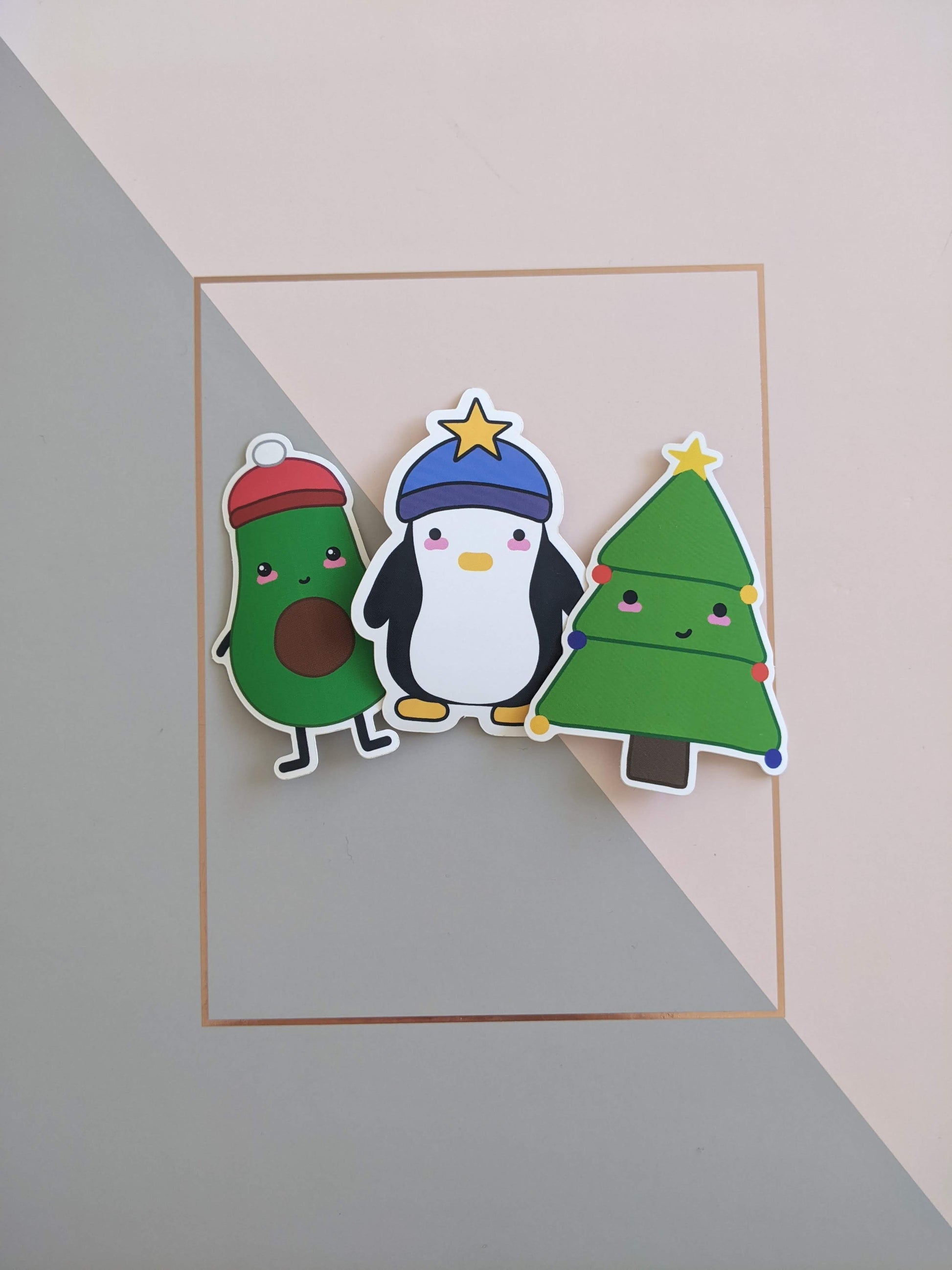 3 stickers on a pink and grey background. On the left is a halved avocado with a smiley face and blushed cheeks, wearing a red beanie hat with a white pompom on top. In the centre is a chunky penguin with a blank expression and blushed cheeks, wearing a blue beanie hat with a yellow star on top. On the right is a Christmas tree with a cheeky smile and blushed cheeks, he has 6 baubles, 2 red, 2 royal blue and 2 yellow, as well as a yellow star at the top. 