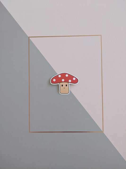 A mushroom sticker on a pink and grey background. She has a beige stalk with a little smiley face and a red top with red spots inspired by fly agaric mushrooms.